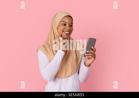 Jeune femme musulmane noire en hijab souriant ludique tout en tenant un smartphone Banque D'Images