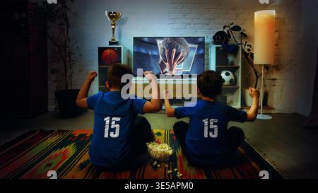 Deux garçons regardant un match de basket-ball à la télévision à la maison célébrant la victoire ensemble. Banque D'Images