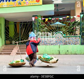 Une femme marche le long d'une rue animée à Hanoi. Elle porte des paniers sur un poteau et porte un chapeau bleu et une veste rouge tandis que les magasins sont à proximité. Banque D'Images