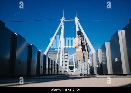 Homme sur le pont piétonnier moderne vérifiant smartphone dans la tenue de mode urbaine d'hiver Banque D'Images