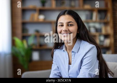Jeune femme indienne souriant en toute confiance sur un canapé confortable dans une maison moderne, détendue et accessible avec un fond d'étagère flou, idéal pour le style de vie et les concepts de travail à distance Banque D'Images