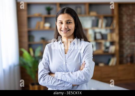 Jeune femme professionnelle confiante debout avec les bras croisés dans un bureau à domicile moderne, souriant à la caméra pour transmettre la détermination, le succès et le leadership dans un environnement de travail de démarrage Banque D'Images