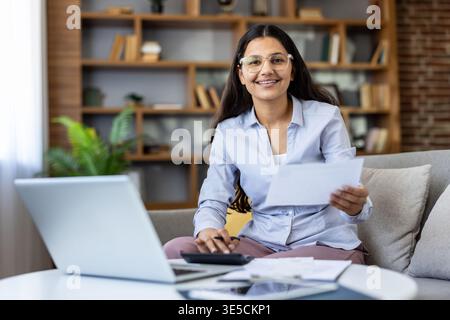 Jeune femme indienne sur un canapé de salon confortable gérant les impôts et les finances personnelles avec ordinateur portable et paperasse, souriant avec confiance tout en budgétisant et planifiant les investissements Banque D'Images