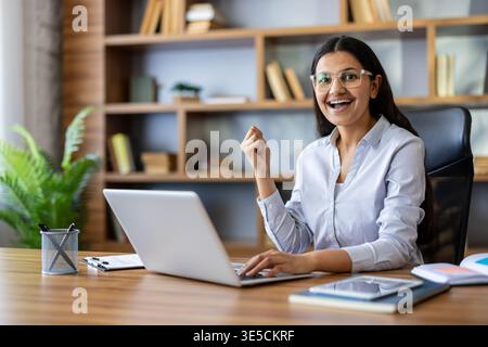 Jeune femme indienne dans les lunettes célèbre le succès à son bureau à domicile, souriant à l'ordinateur portable après avoir atteint des objectifs professionnels tout en travaillant à distance et se sentant confiant et joyeux Banque D'Images