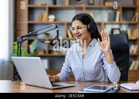 Jeune femme créant un podcast, portant des écouteurs et parlant dans un microphone tout en saluant son ordinateur portable, diffusant depuis un home studio ou un bureau Banque D'Images