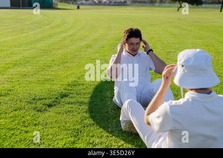 Des coéquipiers masculins effectuant des sit-ups en partenariat sur un terrain tondu dans des vêtements de sport blancs et une montre-bracelet. Athlètes, entraînement, à l'extérieur, herbe, pelouse, ensoleillé, Banque D'Images