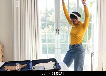 Femme afro-américaine adulte se déplaçant dans une chambre avec valise ouverte, portant un casque, espace de copie. Bagages, écouteurs, denim, moutarde, lunettes de soleil, casua Banque D'Images