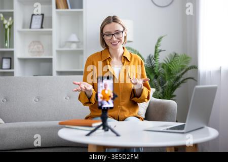Femme enregistrant un blog vidéo ou un cours en ligne à la maison, assise sur un canapé et utilisant un smartphone sur un trépied, créant ainsi du contenu numérique frais pour son public Banque D'Images