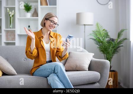 Jeune femme sur le canapé souriant et agitant pendant un appel vidéo sur smartphone depuis son salon, communiquant pour le travail à distance, le chat social ou une réunion en ligne Banque D'Images