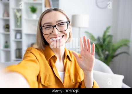 Jeune femme portant des lunettes et une chemise jaune, souriant joyeusement et agitant devant la caméra lors d'une vidéoconférence à la maison, représentant la communication et l'accueil à distance Banque D'Images