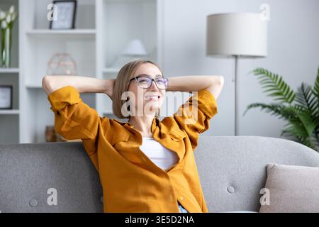 Jeune femme en lunettes et une chemise jaune est assise sur un canapé à la maison, les mains derrière la tête, souriant paisiblement et profitant d'un moment calme et confortable de contemplation détendue Banque D'Images