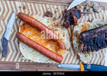 Un sandwich rustique à la saucisse grillée sur du pain grillé avec du bacon carbonisé et des couteaux éparpillés sur une planche à découper en bois à motifs, évoquant backya Banque D'Images