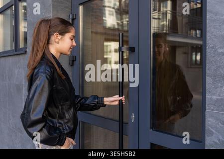 Jeune femme ouvrant la porte du bâtiment moderne. Vue de côté d'une jeune femme dans une veste en cuir noir ouvrant une porte d'entrée en verre dans un urbain moderne Banque D'Images