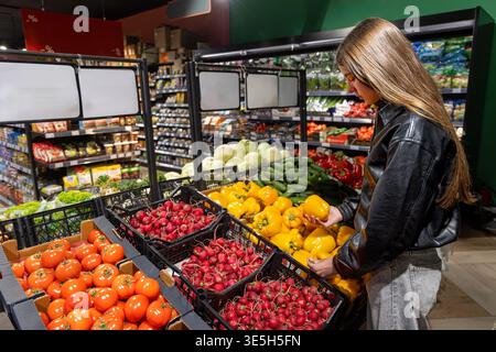 Jeune femme choisissant les poivrons dans l'épicerie. Vue de côté d'une jeune femme sélectionnant des poivrons jaunes frais dans une allée de produits de supermarché remplie Banque D'Images