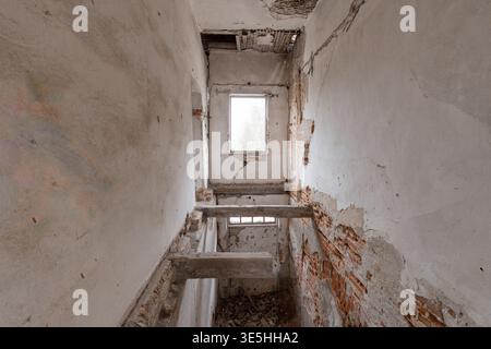Les escaliers en béton qui s'effondrent mènent à travers un couloir étroit où la peinture blanche pelée révèle des briques rouges sous les poutres de plafond endommagées, éclairées par un d mou Banque D'Images