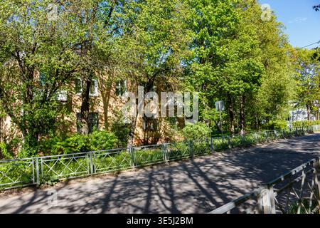 Scène urbaine baignée de soleil : un charmant bâtiment jette un œil à travers un feuillage vert dense, avec de longues ombres dansant sur le trottoir à côté d'une clôture métallique Banque D'Images