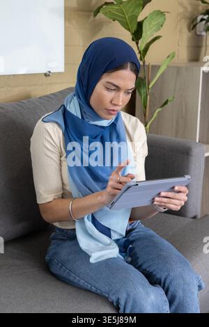 Femme assise sur un canapé gris dans un petit salon de bureau à l'aide d'une tablette et portant un hijab dégradé bleu Banque D'Images