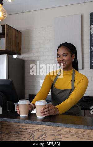 Barista afro-américaine en haut moutarde et tablier gris offrant deux tasses à emporter au comptoir Banque D'Images