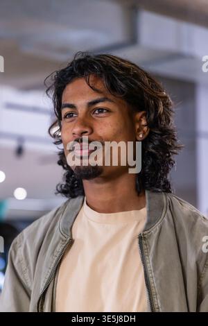 Homme indien dans la vingtaine souriant, regardant à l'intérieur du café portant une veste zippée et un goujon d'oreille gauche Banque D'Images