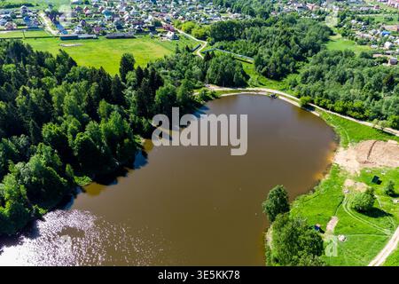 Vue aérienne d'un étang tranquille et trouble niché au milieu de forêts verdoyantes et d'un village lointain. Un chemin rustique longe le bord de l'eau, mettant en valeur rur Banque D'Images