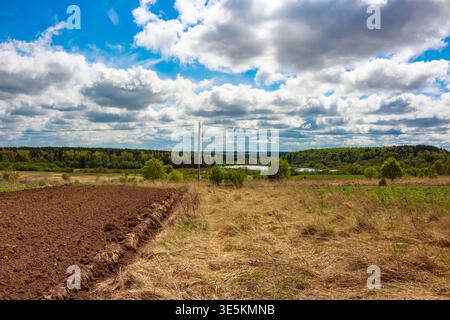 La terre brune fraîchement labourée rencontre un champ d'herbe sèche embrassée par le soleil sous un ciel bleu vif rempli de nuages blancs moelleux, évoquant le renouveau printanier an Banque D'Images