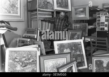 Sloviansk, années 1980, studio de création d'artistes. Photographie d'archives vintage en noir et blanc représentant l'artiste distingué Yuri Grigorievich Savchenko debout gracieusement. Le peintre repose sa main sur un grand support en bois rempli de nombreuses toiles, tandis que plusieurs peintures de paysage magnifiquement encadrées sont dispersées sur le sol au premier plan, capturant l'espace de travail inspirant, le processus artistique authentique de l'ère soviétique et l'histoire culturelle profondément enracinée trouvée dans le Donbass paisible. Banque D'Images