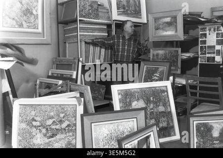 Sloviansk, années 1980, studio de création d'artistes. Photographie d'archives vintage en noir et blanc représentant l'artiste distingué Yuri Grigorievich Savchenko debout gracieusement. Le peintre repose sa main sur un grand support en bois rempli de nombreuses toiles, tandis que plusieurs peintures de paysage magnifiquement encadrées sont dispersées sur le sol au premier plan, capturant l'espace de travail inspirant, le processus artistique authentique de l'ère soviétique et l'histoire culturelle profondément enracinée trouvée dans le Donbass paisible. Banque D'Images