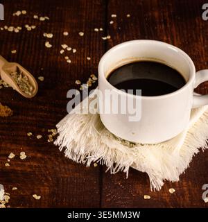 Gros plan de la tasse à café noire sur tissu de lin avec des flocons d'avoine dispersés sur une table en bois rustique, concept de petit déjeuner minimal. Banque D'Images