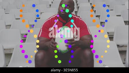 Homme noir assis tenant un ballon de rugby blanc-vert dans des sièges de stade, hexagones verticaux, maillot rouge Banque D'Images