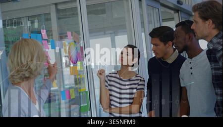 Pointage femme principale guidant l'équipe étudiant le panneau de verre dans le bureau portant cardigan, roue de couleur Banque D'Images