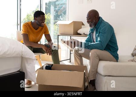 Déballer père et fils afro-américains à côté de la boîte de déménagement à la maison, tenant sneaker et smartphone Banque D'Images