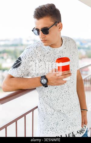 Cool beau hipster homme avec des lunettes de soleil classiques dans un T-shirt blanc à la mode tient le café et se tient sur le balcon Banque D'Images