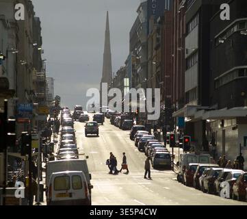 Scène animée de Bath Street avec des piétons traversant une route urbaine ensoleillée, bordée de voitures garées et de grands bâtiments. L'image transmet les déplacements, la ville lif Banque D'Images