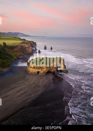 Vue aérienne des rochers des trois Sœurs se dressant majestueusement contre l'océan, avec des sables noirs contrastant avec les falaises vertes, Tongapōrutu, Taranaki R. Banque D'Images