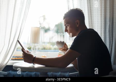 le jeune homme est assis dans un café près de la fenêtre, boit du café et lit un livre Banque D'Images