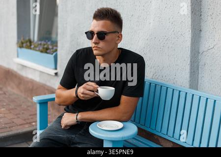 Mode bel homme avec des lunettes de soleil se trouve sur le banc et boit du café Banque D'Images
