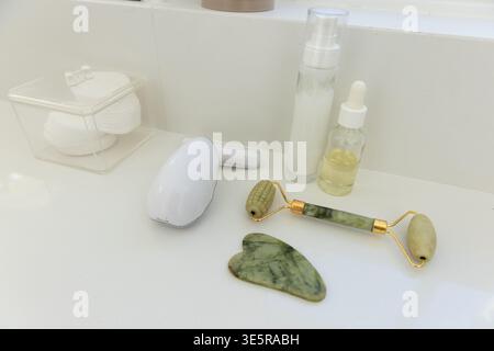 Rouleau de jade et gua Sha reposant sur le comptoir de salle de bain brillant avec compte-gouttes d'huile, pompe de lotion, dispositif Banque D'Images