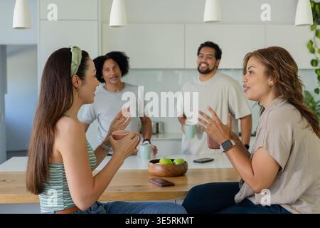 Amis assis sur des tabourets dans la cuisine moderne tenant des tasses près d'un bol de pommes vertes et smartwatch Banque D'Images
