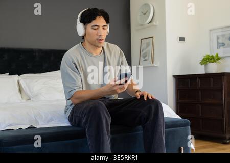 Homme asiatique assis sur le bord du lit portant des écouteurs blancs et tenant un smartphone dans une chambre soignée Banque D'Images