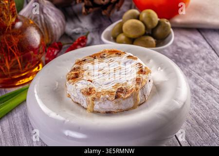 Fromage camembert grillé gourmand sur un plat en céramique blanche. Cette scène d'apéritif rustique présente des olives fraîches, de l'ail, de la tomate et de l'huile infusée de piment Banque D'Images