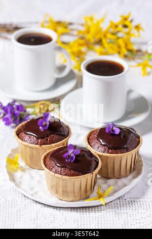 Cupcakes au chocolat garnis de glaçage et de violettes servis avec du café, entourés de fleurs de violette et de forsythia, créant une compos de dessert printanier doux Banque D'Images