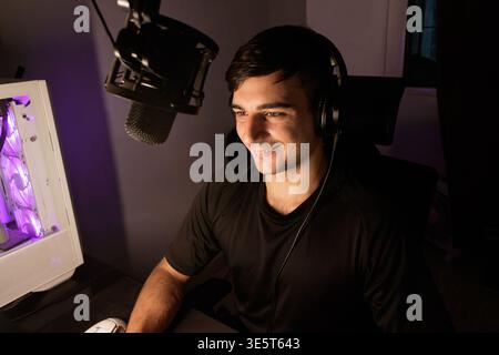 Un jeune homme caucasien heureux est assis à son bureau équipé d'un microphone professionnel, d'écouteurs et d'un pc de jeu. L'éclairage est un mélange de pièce chaleureuse Banque D'Images