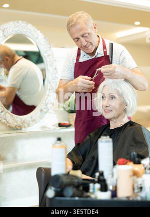 Le maître expérimenté de l'homme senior fait effectue la coupe de cheveux pour la femme mature avec des ciseaux Banque D'Images