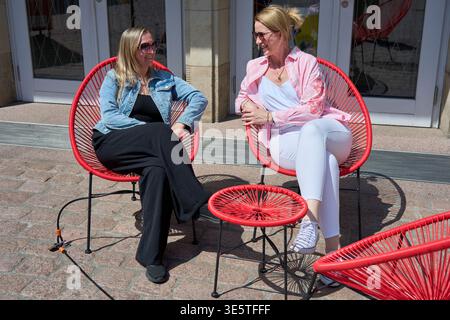 Deux joyeuses femmes d'âge moyen assis dans des chaises rouges Acapulco sur une terrasse ensoleillée, discutant et profitant d'une journée d'été. Style de vie et amitié. Banque D'Images