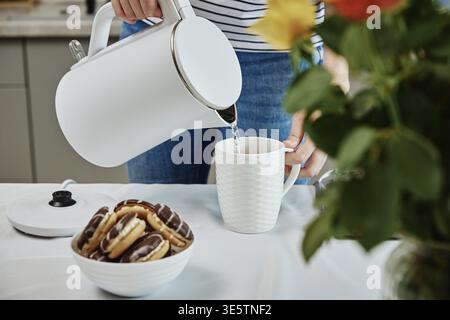 Temps du thé, femme versant de l'eau bouillie de la bouilloire électrique dans la tasse pour préparer le thé, petit déjeuner dans la cuisine Banque D'Images