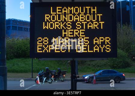 LONDRES, ROYAUME-UNI. 2 avril 2026 : des motocyclistes et une voiture attendent aux feux rouges sous un panneau d'avertissement électronique au rond-point de Leamouth (A1020). La signalisation avertit les automobilistes des travaux publics majeurs et des retards importants prévus entre le 28 mars et le 10 mai 2026. Cette documentation, qui fait partie de l'archive « Newham Portal », capture les frictions liées aux voyages avant Pâques et l'impact de la maintenance à long terme des infrastructures sur les banlieusards de l'est de Londres. Crédit : xiu bao/Alamy Live News Banque D'Images