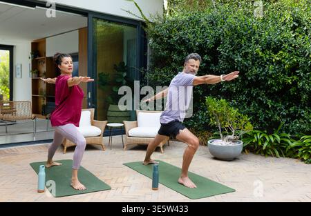 Couple diversifié pratiquant le yoga debout dans des vêtements de gymnastique sur le patio avec des tapis verts et des bouteilles d'eau Banque D'Images