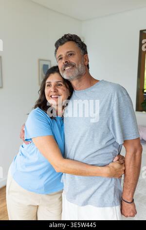 Couple embrassant et souriant dans une chambre lumineuse près de l'art encadré, lit, plante en pot, bracelet Banque D'Images