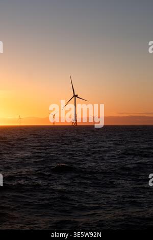 Rangée d'éoliennes offshore silhouettée contre un coucher de soleil orange et or vif sur la mer du Nord, avec des collines côtières visibles au-delà. Banque D'Images