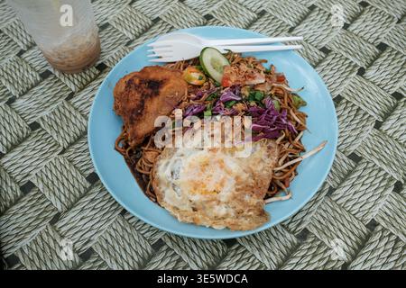 Nouilles fraîches sautées garnies d'oeufs, de concombre et de chou violet. Aile de poulet frite est assis à côté du plat. La fourche en plastique repose sur le bord du Banque D'Images